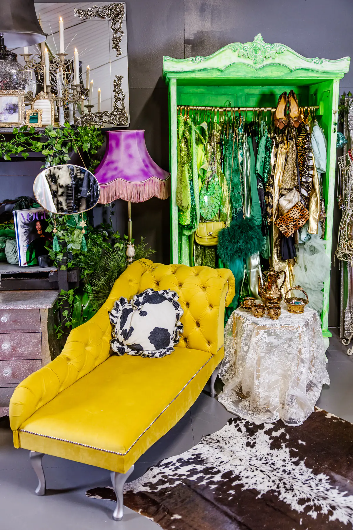 The styling room empty and waiting: a yellow velvet chaise, a bright green painted wardrobe hung with green and gold pieces, a pink fringed lampshade and a cowhide rug.