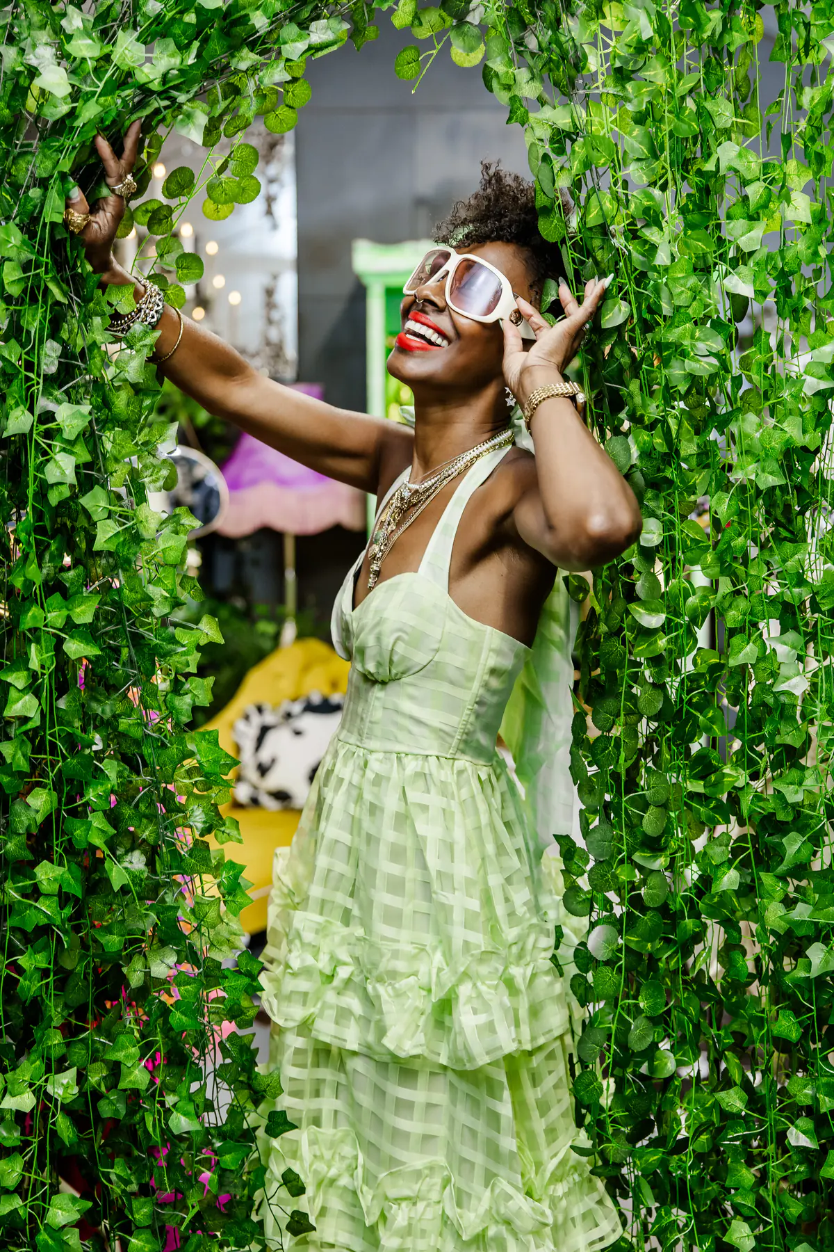Deborah laughing in a lime green tiered dress and white sunglasses, standing against a wall of trailing ivy.