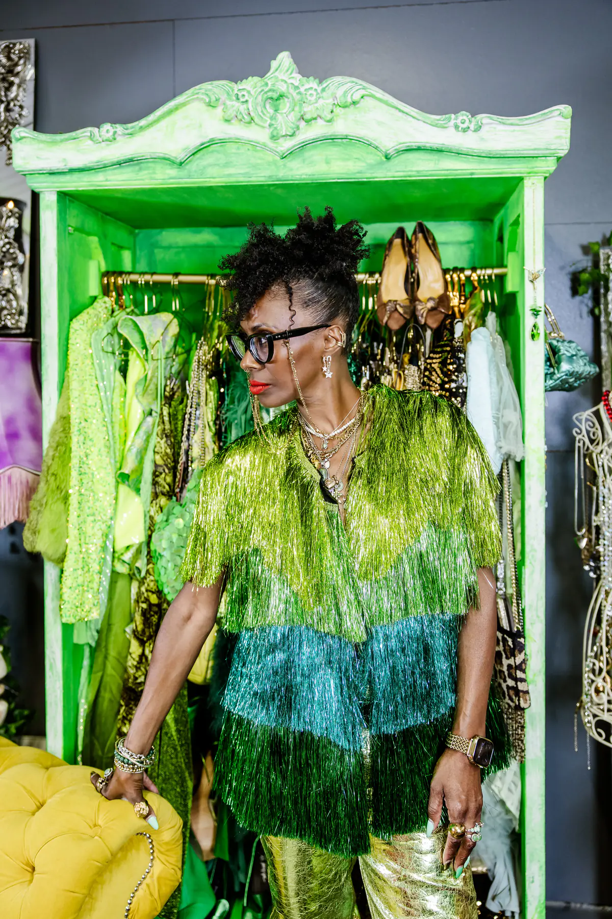 Deborah standing at the green wardrobe in a fringed jacket layered green to teal, with gold metallic trousers.