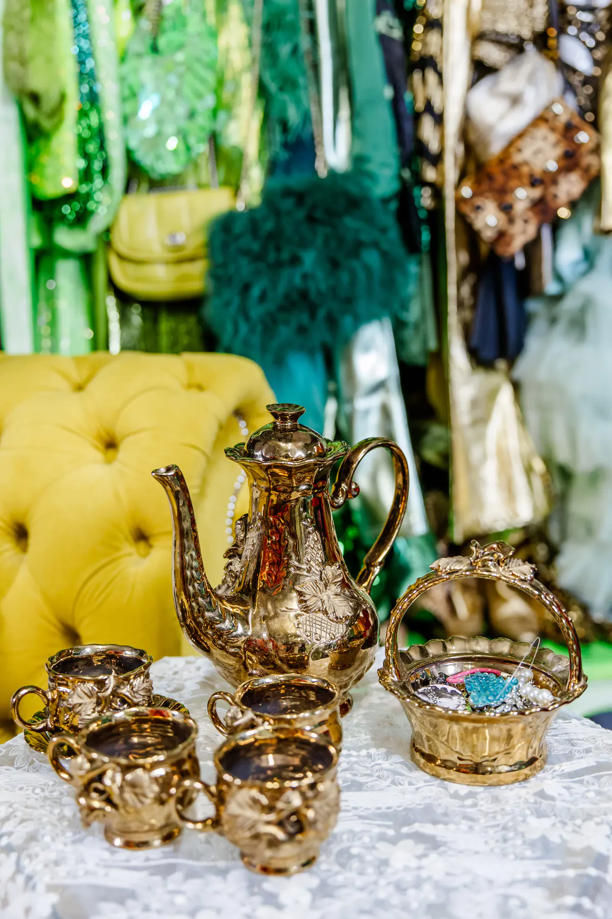 A gold coffee pot, small gold cups and a gilded basket arranged on lace, the yellow chaise behind them.